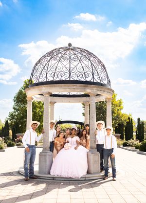 Quinceañera in a pink ball gown poses with court members in cowboy hats and boots beneath an ornate gazebo on a sunny day.
