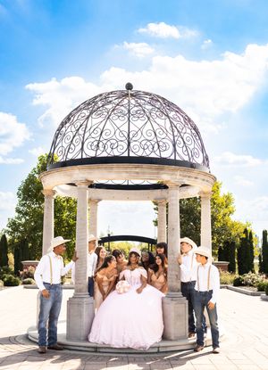 Quinceañera in a pink ball gown poses with her court and men in cowboy hats beneath an ornate stone gazebo on a sunny day.