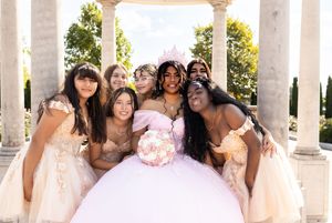 Smiling quinceañera in a pink ball gown and tiara poses with her court in cream dresses beneath a stone gazebo with columns.