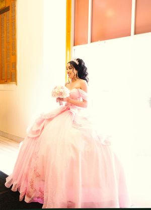 Young woman in a flowing pink quinceañera ball gown with floral embroidery, tiara and bouquet of pink roses, posing beside a sunlit window.