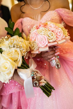 Woman in a sparkly pink gown holds a bridal bouquet of cream and pink roses with feathers, baby's breath, and ribbon-wrapped photo charms.