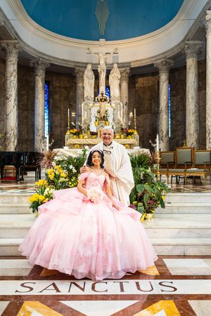 Quinceañera in a pink ball gown holding a bouquet poses with a priest on the altar steps of a marble church decorated with yellow flowers.