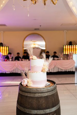 Two-tier white and blush wedding cake with roses and pampas grass on a wooden barrel in an elegant reception hall.