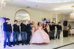 Quinceañera in a pink ball gown poses with family and friends in dark outfits raising glasses in a toast inside an elegant banquet hall.