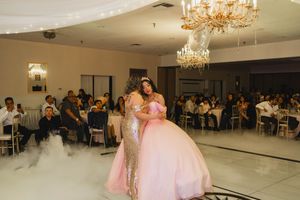 Two women in pink and gold dresses embrace on a dance floor surrounded by low fog, with guests seated at tables beneath crystal chandeliers.