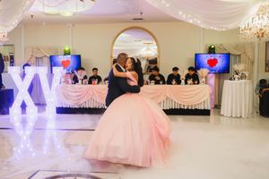 Quinceañera in a pink ball gown dances with a man in a suit on a banquet hall floor beside glowing XV letters and a decorated head table.