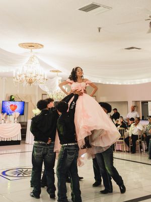 Young woman in a pink quinceañera gown is lifted high by a group of young men during a celebration in a chandelier-lit banquet hall.