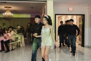 A young woman in a sparkling mint dress and tiara dances with a man in dark clothing at a quinceañera celebration while guests watch.