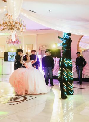 Quinceañera in a pink ball gown sits on the dance floor as an LED-suited stilt performer and guests watch in a chandelier-lit banquet hall.