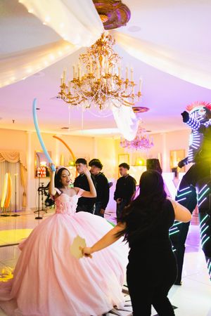 Girl in a pink quinceañera ball gown dances with a blue balloon sword beside a glowing LED robot performer under a crystal chandelier.