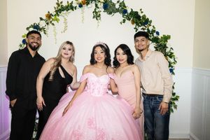 Quinceañera in a pink ball gown and tiara poses with two women and two men in front of a floral greenery arch.