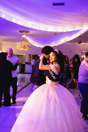 A quinceañera in a glowing pink ball gown and sash dances with a partner under draped lights and a chandelier at a festive reception.