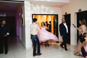 A young woman in a pink quinceañera ball gown poses in a doorway while guests in formal attire watch and take photos at the celebration.