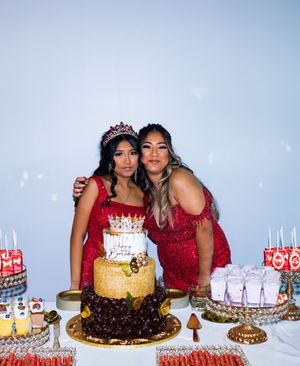 Two women in red sequin dresses, one wearing a tiara, pose behind a tiered birthday cake and a decorated dessert table.