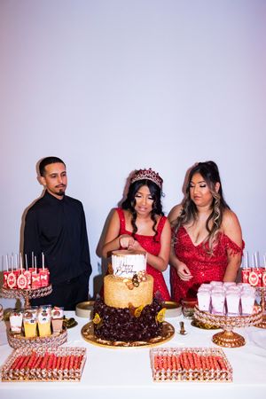 Quinceañera in a red dress and tiara cuts her birthday cake beside two guests at a dessert table with treats.