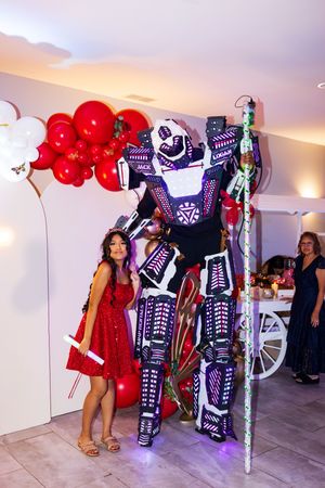 A smiling girl in a red sequined dress poses with a tall LED-lit robot performer beside red and white party balloons.