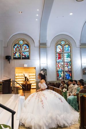 Quinceañera in a voluminous white embroidered ball gown stands near the altar of a church with stained glass windows as guests watch.