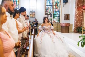 Young woman in a white and pink floral quinceañera ball gown and tiara smiles in a church as family members watch from the pews.