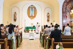 Wedding guests stand in pews decorated with greenery and flowers as a priest leads a Catholic ceremony before an altar with a Virgin Mary im