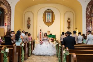 Quinceañera in a white tulle gown kneels at the church altar with a priest while family and guests watch from the pews.
