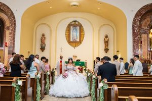 Quinceañera in a white ball gown stands at the altar of a church, surrounded by family and guests in pews decorated with green ribbons.