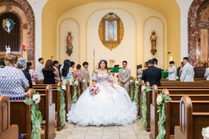 Quinceañera in a sparkling white ball gown holding a bouquet stands in a church aisle lined with floral pews and guests.