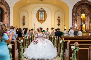 Quinceañera in a sparkling white ballgown stands in a church aisle lined with floral pew decorations as guests watch from the pews.
