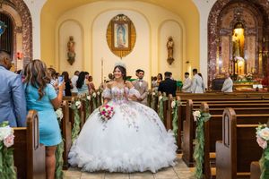 Quinceañera in a floral-embellished white ball gown stands in the church aisle with a bouquet as guests fill the pews.