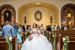 Quinceañera in a floral-embellished white gown walks down the church aisle lined with pews decorated with greenery and white flowers.