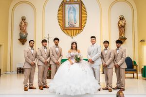 Quinceañera in a white ball gown holds a bouquet while six chambelanes in tan suits pose in front of the Guadalupe image in the church.