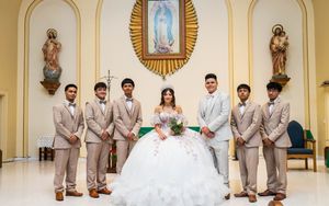 Quinceañera in a layered white ball gown and tiara poses with her seven chambelanes in tan suits inside a church sanctuary.