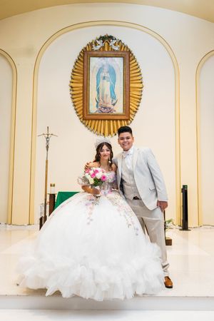 Quinceañera in a white floral-embroidered ball gown holds a pink bouquet and poses with a young man in a gray suit before a church altar.