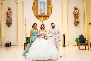 Quinceañera in a white ball gown with bouquet poses with a woman in teal and a man in a gray suit inside a church with religious statues.