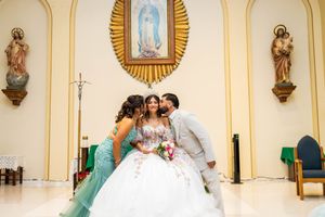 Quinceañera in a white ball gown poses at a church altar while a girl in a teal dress and a man in a light suit kiss her cheeks.