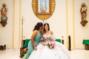 A quinceañera in a white ball gown with a tiara and floral bouquet is kissed on the cheek by a woman in a teal dress inside a church.