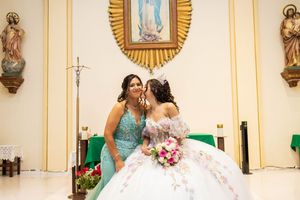 Two young women embrace in a church, one in a white floral quinceañera gown holding a pink bouquet beside the altar.