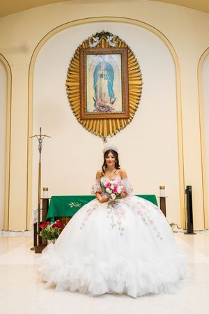 Quinceañera in a white floral ball gown holds a pink bouquet while seated before a Virgin of Guadalupe image in a church.