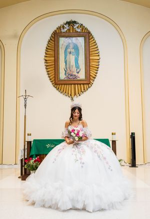 Quinceañera in a white ball gown holds a pink bouquet before an altar beneath a gold-framed Our Lady of Guadalupe image.