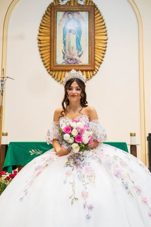 Smiling quinceañera in a silver tiara and white gown holds a pink and white rose bouquet while seated at a church altar.
