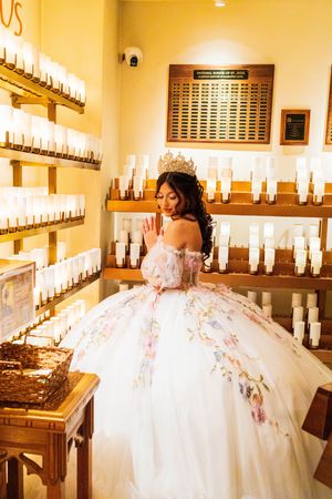 Quinceañera in a white floral ball gown and tiara poses beside shelves of lit votive candles in a warm church prayer room.