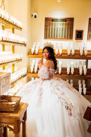 Quinceañera in a floral embroidered ivory ball gown and tiara waves beside rows of votive candles in a warm chapel setting.