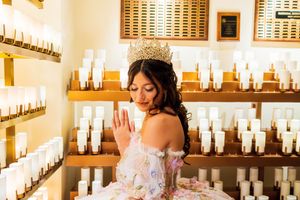 Quinceañera wearing a tiara and a floral-embroidered off-the-shoulder gown raises her hand beside shelves of lit prayer candles.