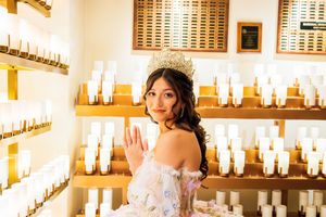 Quinceañera in a tiara and off-the-shoulder floral gown poses beside rows of white votive candles in a church chapel.