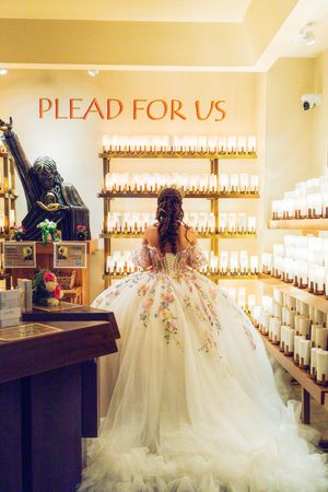 Quinceañera in a floral embroidered ball gown stands before glowing prayer candles and a statue beneath a Plead for Us sign.
