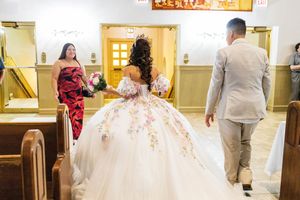 Quinceañera in a floral-embellished white ball gown walks into a church beside a man in a tan suit as a woman holding flowers watches.