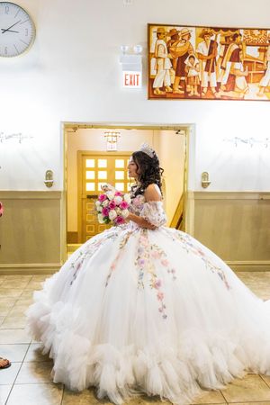 Quinceañera in a floral-embroidered white tulle gown and tiara holds a pink bouquet beside a doorway and a colorful mural.