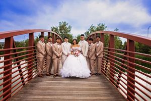 Quinceañera in a white ball gown holding a bouquet poses on a wooden bridge surrounded by seven escorts in tan suits.