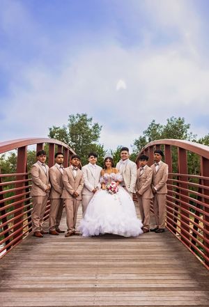 Quinceañera in a white tulle ball gown holds a bouquet on a wooden bridge, surrounded by seven young men in tan suits.