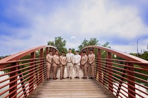 Seven chambelanes in tan suits stand in a row on a red-railed wooden bridge under a blue sky with clouds.