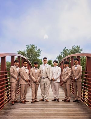 Seven young men in matching tan suits and bow ties pose together on a red arched wooden bridge under a partly cloudy sky.